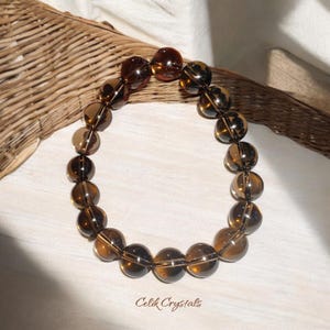 Peut inclure: Un bracelet en pierres précieuses de quartz fumé brun avec des perles rondes. Le bracelet est fait de pierres naturelles et a une couleur brun foncé avec une légère teinte dorée. Les perles sont lisses et polies, et le bracelet est enfilé sur un cordon élastique transparent.