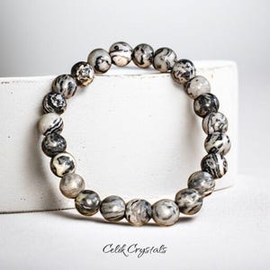May include: A beaded bracelet featuring round, patterned beads in shades of gray, black, and white. The bracelet is displayed on a white surface. The text "Celik Crystals" is visible at the bottom.
