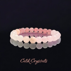 May include: A pink and light pink gemstone bracelet with round beads. The bracelet is on a black background and reflects in a mirror.
