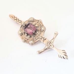 May include: A silver-coloured brooch in the shape of a sword. The brooch features a central, rectangular, purple gemstone surrounded by ornate detailing. The sword's hilt has a Celtic knot design and a leaf-shaped tip.