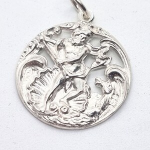 Puede incluir: Colgante de plata con un relieve detallado de una figura mitológica, posiblemente Poseidón, montando un caballito de mar entre olas y una concha. El colgante es circular con un borde calado ornamentado.