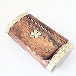 Puede incluir: Una caja rectangular de madera con un emblema de trébol de cuatro hojas. La caja tiene un acabado marrón oscuro con bordes de metal plateado. El trébol es de color dorado. La caja probablemente se utiliza para guardar objetos pequeños.