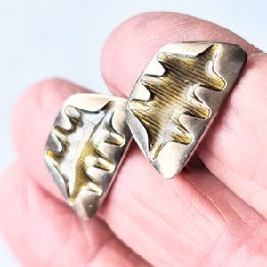 Peut inclure: Une paire de boutons de manchette argentés avec un motif de feuille dorée. Les boutons de manchette sont de forme rectangulaire avec un motif organique texturé au centre. Le motif est visible sur un fond clair.