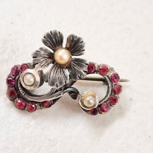 Op de afbeelding: Zilverkleurige broche met een bloem met een parel in het midden en een gebogen ontwerp versierd met kleine, rode edelstenen en extra parels. De broche heeft een speldsluiting aan de achterkant.