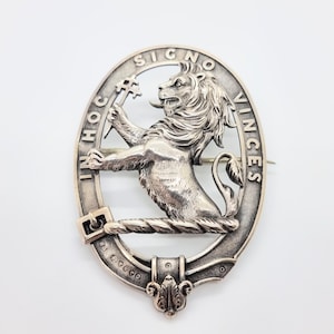 以下が含まれることがあります： 銀色の楕円形のブローチ。鍵を持つライオンが描かれ、ラテン語の「IN HOC SIGNO VINCES」の文字が周囲に刻まれています。ブローチはピン留め式で、装飾的な要素があります。