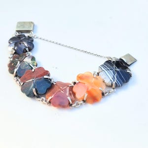 Puede incluir: Un brazalete de cadena de plata con una serie de abalorios en forma de trébol hechos de diferentes tipos de ágata. Los abalorios son de color negro, verde, rojo, naranja y blanco.