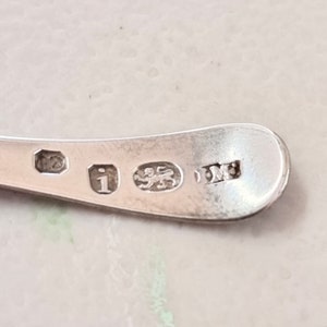 May include: Close-up of the back of a silver spoon with hallmarks. The hallmarks include a lion, a letter 'I', and a maker's mark.