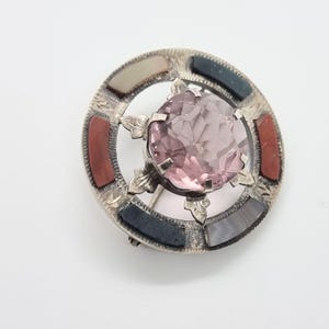 May include: A silver brooch with a pink glass center surrounded by a ring of colorful stones. The stones are red, black, white, and gray.