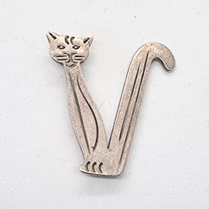 Peut inclure: Une broche argentée en forme de chat. La broche représente un chat stylisé avec une longue queue incurvée formant un V. Le visage du chat présente des détails gravés et le corps des lignes gravées. La broche est probablement en métal.