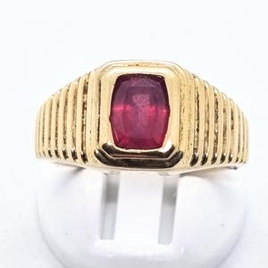 Op de afbeelding: Een gouden ring met een rechthoekige, dieprode edelsteen in het midden. De ring heeft een geribbeld ontwerp op de band en een vierkante zetting rond de edelsteen. De ring staat tegen een witte achtergrond.
