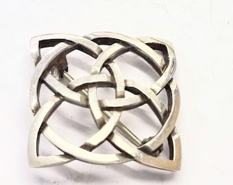 May include: A silver-toned brooch with a Celtic knot design. The brooch features an intricate interwoven pattern, with a pin on the back for secure fastening. The metal has a slightly aged appearance, adding to its vintage charm. The brooch is square-shaped.