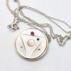 May include: A silver pendant necklace with a circular pendant. The pendant features a triangle design with a pink, black, and red gemstone. The necklace chain is silver and has a lobster clasp.