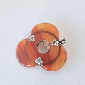 May include: A vintage brooch with a clover-like design. The brooch features three orange and brown agate circles, connected by silver-toned floral accents. A silver pin and clasp are visible on the back. The brooch is a unique piece of jewelry.