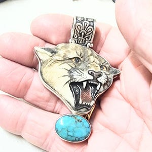 Puede incluir: Un colgante de plata con una cabeza de puma detallada con la boca abierta, revelando dientes y un interior rojo. Debajo del puma hay una piedra turquesa. El colgante se sostiene en una mano.