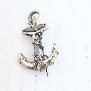 May include: A silver-toned metal brooch shaped like an anchor, featuring an inlaid dark grey iridescent material on the crossbar and shaft. The anchor is wrapped in a twisted metal rope design with engraved detailing on the metal surfaces.