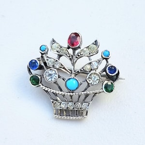 May include: A silver-toned brooch in the shape of a basket filled with colorful gemstones. The brooch features a red ruby, turquoise, clear crystals, emerald green, and sapphire blue stones. The basket design has a detailed woven pattern.