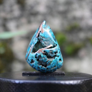 Puede incluir: Una piedra preciosa turquesa en forma de lágrima con inclusiones de cobre. La piedra preciosa tiene una superficie rugosa y texturizada y se muestra en un soporte negro.
