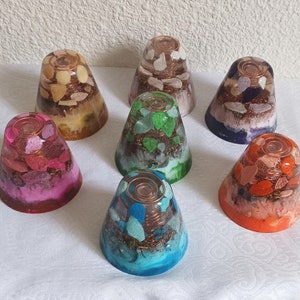 Peut inclure: Sept pyramides d'orgonite de guérison cristalline colorées, multicouches, remplies de résine, avec du fil de cuivre et divers cristaux. Les pyramides sont disposées en cercle sur une surface blanche.