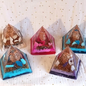 Op de afbeelding: Vijf heldere harspiramides met koperen draadspiralen erin. Elke piramide bevat kristallen en glitter in verschillende kleuren. De piramides zijn gerangschikt op een witte stoffen achtergrond.