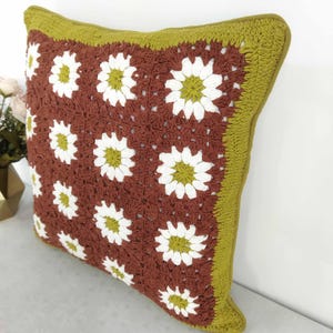 May include: A square crochet pillow featuring a daisy pattern. The pillow has a brown center with white and yellow daisies, a green border, and a green trim. The pillow is approximately 16 inches square.