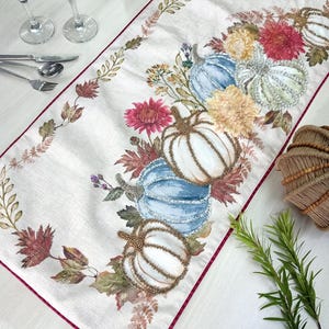 Könnte beinhalten: Cremefarbener Tischläufer mit burgunderrotem Rand, bestickt mit weißen, blauen und grünen Kürbissen, akzentuiert mit goldenen Perlen und farbenfrohen Blumenarrangements. Der Läufer ist mit Herbstblättern verziert.