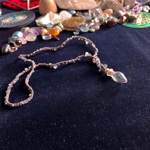 Può includere: Una collana all'uncinetto marrone con piccole perline trasparenti intrecciate lungo il cordino. Una minuscola fiala di vetro trasparente a forma di goccia con un tappo di sughero è fissata all'estremità del cordino.