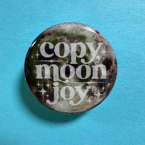 May include: A round button with a moon design and the words "copy, moon, joy" in white. The button has a glossy finish and is set against a light blue background.