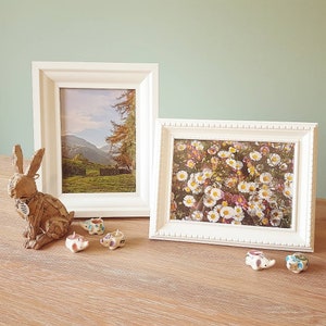 May include: Two white picture frames with different landscape photos. The frame on the left has a photo of a mountain range with trees in the foreground. The frame on the right has a photo of a field of white flowers. There are five small ceramic elephants on a wooden table.