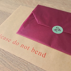May include: A brown paper envelope with the text "Please do not bend" printed in red. A burgundy envelope is on top of the brown envelope with a green sticker with a floral design.