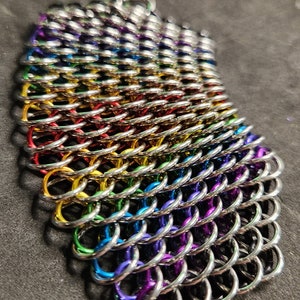 Puede incluir: Un brazalete de malla de cadena arcoíris hecho de anillos plateados con un acabado colorido. El brazalete está formado por múltiples filas de anillos, cada fila de un color diferente del arcoíris.