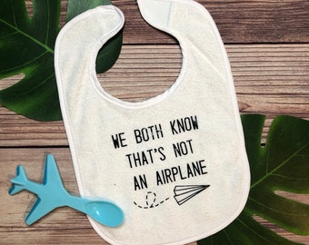 May include: White baby bib with the text "WE BOTH KNOW THAT'S NOT AN AIRPLANE" in black. A blue airplane and spoon are next to the bib. The bib has a velcro closure. The background is a wooden surface with green leaves.