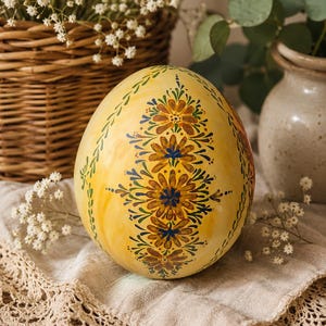 May include: A decorative egg with a smooth yellow finish, featuring a vertical hand-painted pattern of orange and blue flowers with green leafy accents. The egg displays intricate floral motifs arranged in a central column, showcasing detailed brushwork and a glossy surface texture that reflects light across its rounded, symmetrical form.