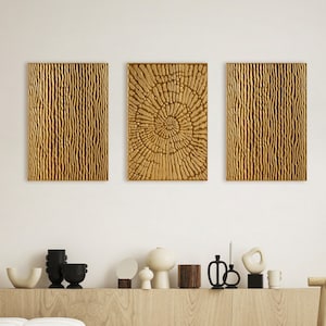 Könnte beinhalten: Drei abstrakte Wandbilder aus Holz in einem Wohnzimmer. Das mittlere Stück zeigt ein kreisförmiges Muster, während die beiden seitlichen Stücke ein vertikales Holzmaserungsmuster aufweisen. Die Kunstwerke sind an einer weißen Wand über einem hellen Holzregal mit Dekorationsgegenständen angebracht.