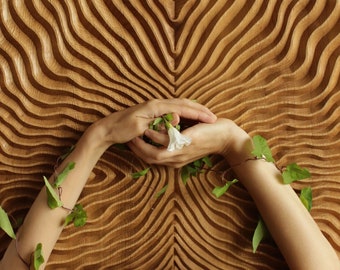 Puede incluir: Un panel de pared rectangular de madera con un patrón ondulado tallado y simétrico. La superficie de madera marrón claro muestra surcos verticales profundos y ondulantes que crean un efecto texturizado y rítmico.