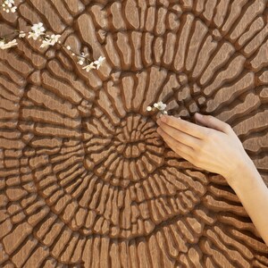 Pode incluir: Um close-up de uma peça de arte de parede de madeira com um padrão circular esculpido na superfície. A madeira é de uma cor castanho rica e o padrão é intrincado e detalhado. Uma mão se estende para tocar a arte de parede, e um pequeno ramo de flores brancas repousa sobre a superfície.