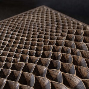 May include: Close-up of a wooden surface with a repeating geometric pattern. The pattern consists of small, raised, pyramid-shaped blocks arranged in a grid. The wood has a dark brown color with visible grain.