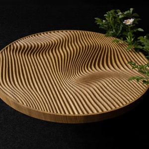 May include: A round wooden serving tray with a wavy, carved pattern. The tray is made of light brown wood and has a natural finish. The tray is sitting on a black surface.