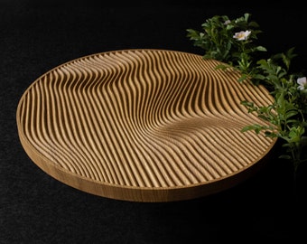 May include: A circular wooden tray featuring a carved surface with deep, undulating wave-like grooves. The light brown wood grain is visible across the textured pattern, which creates a rhythmic, flowing effect across the entire round object.