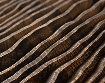 May include: A dark brown wooden surface featuring a series of parallel, undulating, wave-like ridges. The wood grain is visible across the textured, carved peaks and valleys, creating a rhythmic pattern of light and shadow across the material.