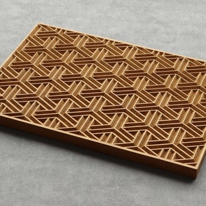 May include: A rectangular wooden tray with a geometric pattern. The tray is made of light brown wood and features a repeating design of interlocking shapes. The design creates a three-dimensional effect. The tray is on a grey surface.