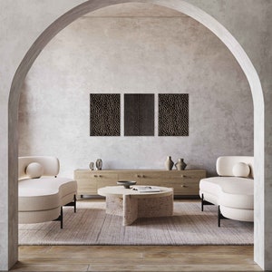 May include: A modern living room scene featuring a neutral color palette. Two cream-colored curved armchairs flank a round coffee table. Above, three textured wall art pieces hang on the wall. A wooden cabinet and vases complete the decor.