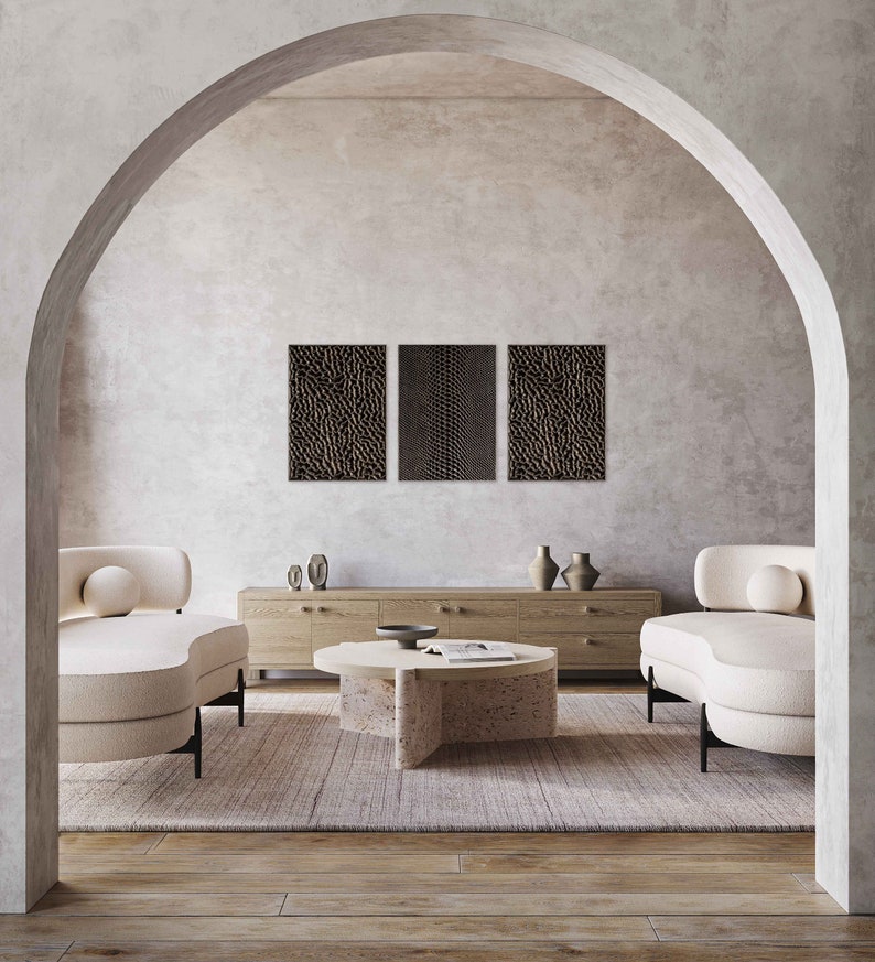 May include: A modern living room scene featuring a neutral color palette. Two cream-colored curved armchairs flank a round coffee table. Above, three textured wall art pieces hang on the wall. A wooden cabinet and vases complete the decor.