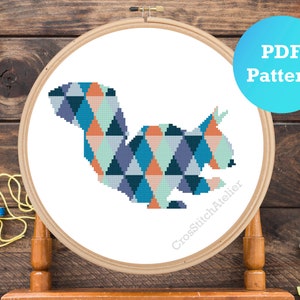 May include: A cross stitch pattern of a geometric squirrel in shades of blue, green, orange, and pink. The pattern is displayed in a wooden embroidery hoop. The text "PDF Pattern" is displayed in a blue circle.