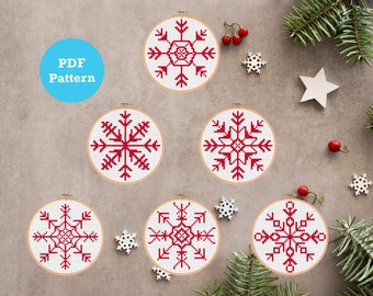 Puede incluir: Seis patrones de bordado de copo de nieve rojo y blanco en aros de madera redondos. Los aros están dispuestos en semicírculo sobre un fondo gris. El texto "PDF Pattern" está en un círculo azul en la esquina superior izquierda de la imagen.
