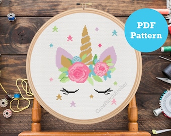 May include: A cross stitch pattern featuring a unicorn head with a gold horn, pink flowers, and eyelashes. The pattern is in a white embroidery hoop on a wooden background. The text "PDF Pattern" is on a blue circle.