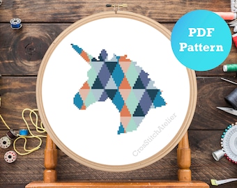 Może przedstawiać: Geometriczny wzór haftu krzyżykowego przedstawiający głowę jednorożca w odcieniach niebieskiego, turkusowego, pomarańczowego i białego. Wzór znajduje się w drewnianej obręczy do haftu, a tekst "PDF Pattern" znajduje się w niebieskim kółku.