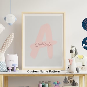 May include: A framed cross-stitch pattern featuring a large pink letter "A" and the name "Adele" in a cursive font. The words "Custom Name Pattern" are below the frame. The image is displayed in a child's room setting.