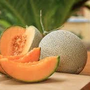 May include: A whole cantaloupe and a halved cantaloupe with several slices on a wooden cutting board. The cantaloupe has a textured, light green rind and bright orange flesh. The background is blurred, showing green foliage.