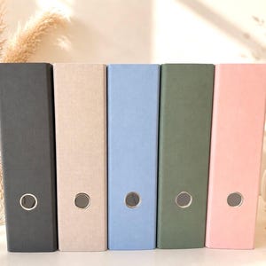 Op de afbeelding: Een set van vijf verticale ringbanden die op een rij staan, met een textuur die lijkt op stof in antracietgrijs, beige, lichtblauw, saliegroen en zachtroze. Elke ringband is voorzien van een rond zilverkleurig metalen vingergat aan de onderkant.