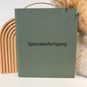 Könnte beinhalten: Ein lindgrünes Buch mit dem Text "Spezialanfertigung" in Schwarz. Das Buch hat einen goldenen Griff und steht neben einem dekorativen Holzbogen und Trockenblumen.